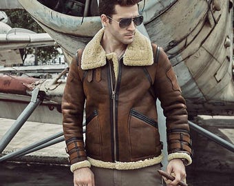 May include: A brown leather flight jacket featuring a thick beige shearling collar, cuffs, and hem. The jacket includes black leather trim along the zipper and pockets, with adjustable buckled straps on the sleeves and a full-length front zipper.