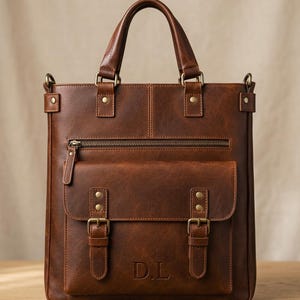 May include: A brown leather vertical tote bag featuring two top handles, a zippered front pocket, and a larger flap pocket secured with two brass-toned buckles. The front flap is embossed with the letters D.L. and includes metal rivets.