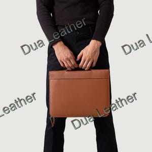 May include: A brown leather briefcase with a handle and zipper closure. The briefcase is rectangular and appears to be made of high-quality leather. The words "Dua Leather" are printed on the image.