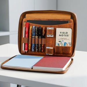 Könnte beinhalten: Eine hellbraune Leder-Reißverschlussmappe mit einem Innenfach, das mehrere elastische Stiftschlaufen für verschiedene Schreibgeräte, ein Fach für ein Field Notes Notizbuch sowie zwei flache Notizbücher in Hellblau und Rot enthält.