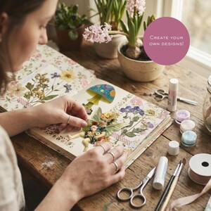 May include: A scrapbook featuring pressed flowers and a fairy illustration with a mushroom, with craft supplies including scissors, glue sticks, and glitter pots on a wooden table. A pink circle contains the text: CREATE YOUR OWN DESIGNS!