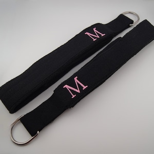 May include: Two black fabric straps with silver metal rings. Each strap has a pink embroidered letter "M". The straps are likely for a bag or luggage.