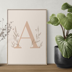 Puede incluir: Un cuadro enmarcado con la letra "A" en un tono beige suave, adornado con ilustraciones botánicas delicadas. Un jarrón blanco con ramas secas y una planta en maceta con hojas verdes grandes completan la decoración.