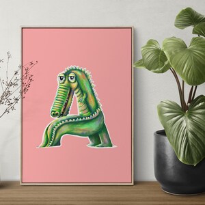 Puede incluir: Impresión enmarcada que presenta una ilustración de un cocodrilo verde con forma de la letra "A" sobre un fondo rosa. La obra de arte se exhibe sobre una superficie de madera, con una planta y un jarrón en el fondo.