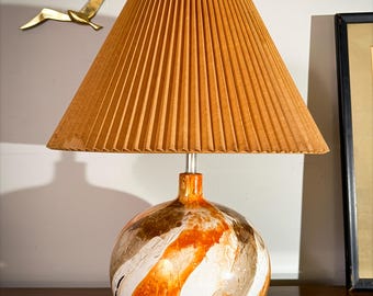May include: A decorative table lamp with a pleated orange lampshade. The base is a round, textured globe with orange, white, and brown swirled patterns. The lamp is on a wooden surface, with gold bird sculptures on the wall behind it.