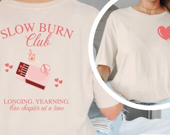 May include: Cream-colored t-shirt with a pink heart on the front and a graphic on the back. The back graphic features the text "SLOW BURN Club" above a matchbox illustration and the words "LONGING. YEARNING. One chapter at a time."