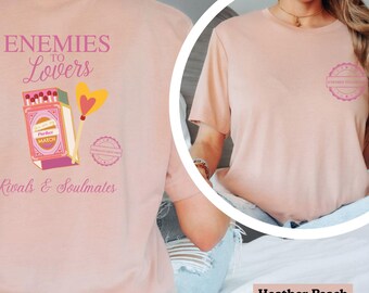 May include: Heather peach-colored t-shirt with the text "Enemies to Lovers" and "Rivals & Soulmates." The design includes a matchbox and a heart-shaped wand. A small circular design is on the right chest. The shirt is a comfortable, casual style.