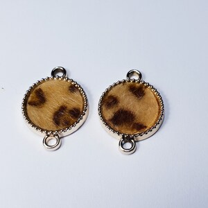 May include: Two gold-toned metal circle charms with a brown and tan leopard print design. Each charm has two loops for attaching to jewelry.