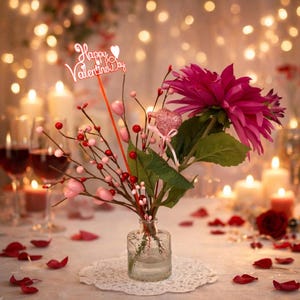 May include: A Valentine's Day centerpiece featuring a clear glass vase with a floral arrangement. The arrangement includes a large pink flower, pink heart-shaped ornaments, and red berries. A sign that says "Happy Valentine's Day" is in the arrangement.