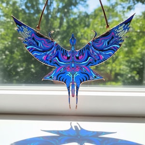 May include: A stained glass suncatcher shaped like a bird with wings spread wide. The piece features intricate swirling patterns in shades of vibrant blue, light blue, and magenta, all outlined in thin copper foil with a hanging wire attached.