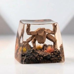 Op de afbeelding: Een heldere, rechthoekige hars keycap met een bruine, gestileerde figuur van een wezen. De keycap heeft een gedetailleerd landschap met bruine rotsen en kleurrijke planten. De figuur lijkt de bovenkant van de keycap omhoog te houden.