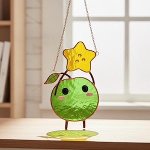 May include: A stained glass suncatcher featuring a round, bright green fruit character with black eyes and pink cheeks, topped with a yellow star and a small green leaf. The piece is suspended by a thin metal chain and has copper-toned edges.