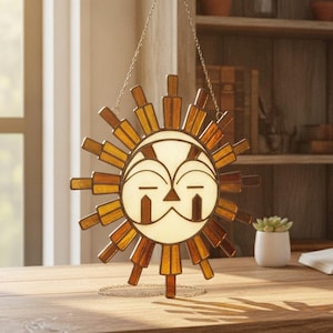May include: A circular sun-shaped stained glass suncatcher featuring a cream-coloured face with stylised brown geometric features and radiating amber-coloured glass rays, suspended by a thin metal chain against a bright indoor setting.