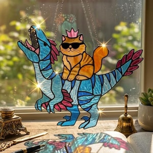 May include: Stained glass suncatcher featuring a cat wearing sunglasses and a crown riding a blue and pink dinosaur. The cat has orange and brown stripes. The dinosaur has blue and pink accents. The suncatcher hangs from a silver chain.