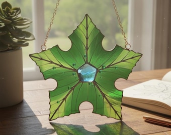 May include: A star-shaped stained glass suncatcher featuring vibrant green leaf-patterned glass panels with dark metal soldering. A single light blue pentagonal glass gem is centered in the middle, accented by small green glass beads along the veins.