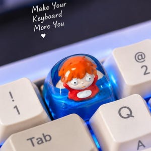 May include: A custom artisan keycap featuring a small, orange-haired character figurine encased in translucent blue resin, positioned on a white mechanical keyboard. The text "Make Your Keyboard More You" with a heart symbol is visible above.