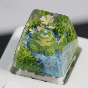 Könnte beinhalten: Eine individuelle Tastenkappe mit einer grünen Kreatur in einer Miniaturlandschaft. Die Tastenkappe hat eine klare, rechteckige Form mit einem Wasserfall und grünem Laub. Rosa und weiße Blumen sind ebenfalls vorhanden.