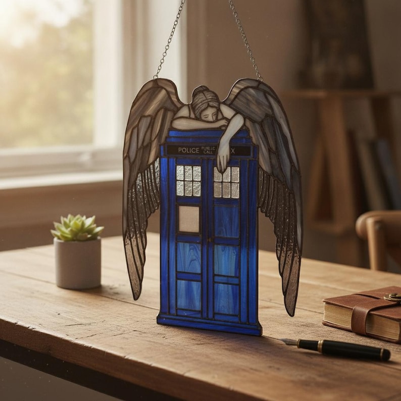 May include: A stained glass suncatcher featuring a blue police box with the text POLICE PUBLIC CALL BOX and a weeping angel figure with large, textured grey wings resting its arms on top of the structure, suspended by a thin metal chain.