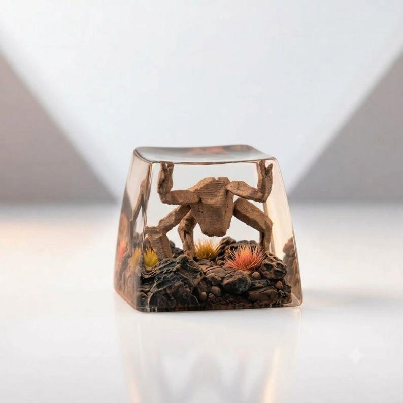 May include: A clear resin mechanical keyboard keycap featuring an encapsulated brown crab figure positioned on a dark, textured rocky base with small yellow and orange tufts of vegetation embedded within the transparent material.