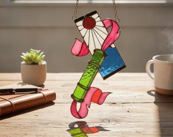 Puede incluir: Un atrapasoles de vidrieras con un tubo central verde similar al bambú, un panel rectangular blanco con un diseño de sol rojo y una sección azul, todo acentuado por cintas rosas fluidas y suspendido por una fina cadena de metal.