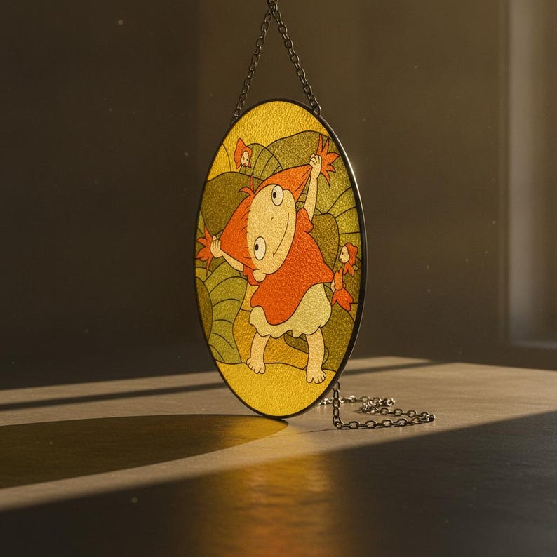 May include: An oval stained glass suncatcher featuring a cartoon character with orange hair and a red shirt, surrounded by green foliage and small orange fish. The piece is framed in a dark metal border and hangs from a silver-toned metal chain.