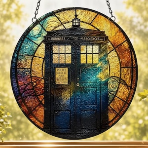 May include: A circular stained glass suncatcher featuring a dark blue police box centered against a swirling background of amber, gold, and deep blue hues. The design includes a small sign with text and a black metal chain for hanging in a window.