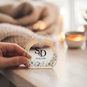 May include: Clear acrylic ring-shaped wedding favour with a diamond-shaped top. The ring features the initials "S&D" and the date "20.05.2026" in black. The ring is decorated with green and white floral accents.