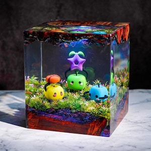 May include: A clear resin cube featuring a miniature scene with cartoon apple figures. The base has a wood-like appearance, and the top has colorful details. The apples are yellow, green, and blue, each with an accessory, set in a grassy area.