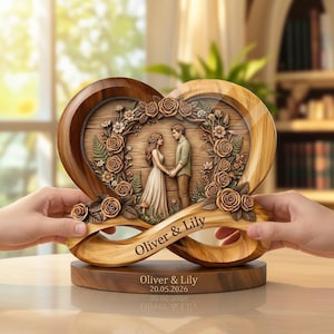 Puede incluir: Escultura de madera en forma de corazón con una pareja tomados de la mano, rodeada de tallas florales. La escultura tiene los nombres "Oliver & Lily" y la fecha "20.05.2026" grabados. La base también es de madera.