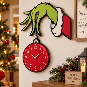 Op de afbeelding: Een wandklok in de vorm van een rode kerstbal die hangt aan een groene hand met een witte boord en een rode mouw. De wijzerplaat is felrood met witte cijfers van 1 tot 12 en goudkleurige uur-, minuut- en secondewijzers.