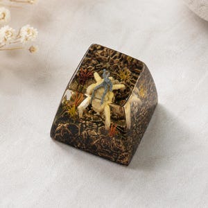 May include: A custom mechanical keyboard keycap featuring a clear resin enclosure with a dark, textured base. Inside, a small, light-colored crab figure with a grey top sits amidst miniature green and orange aquatic plants and rocky terrain.