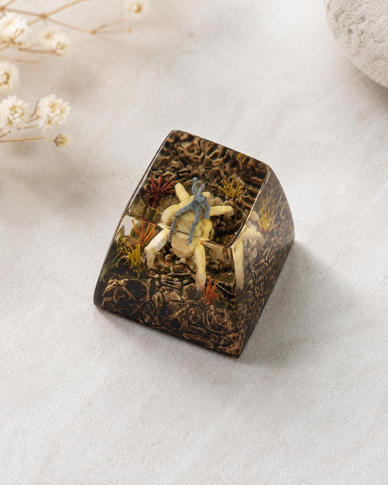 May include: A custom mechanical keyboard keycap featuring a clear resin enclosure with a dark, textured base. Inside, a small, light-colored crab figure with a grey top sits amidst miniature green and orange aquatic plants and rocky terrain.