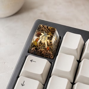 May include: A custom resin mechanical keyboard keycap featuring a miniature beige spider-like creature encased within a textured brown forest floor scene with small orange and yellow plant accents, positioned on a black keyboard with white keys.