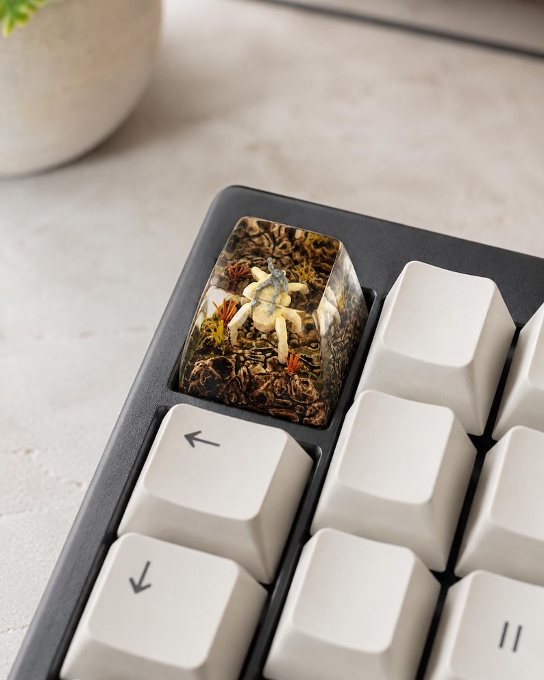 May include: A custom resin mechanical keyboard keycap featuring a miniature beige spider-like creature encased within a textured brown forest floor scene with small orange and yellow plant accents, positioned on a black keyboard with white keys.