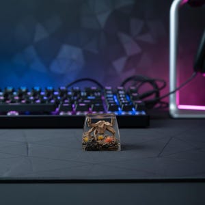 May include: A clear resin mechanical keyboard keycap featuring a miniature brown crab figure embedded inside. The base of the keycap contains dark textured gravel and small orange and yellow tufts of artificial grass surrounding the crab.