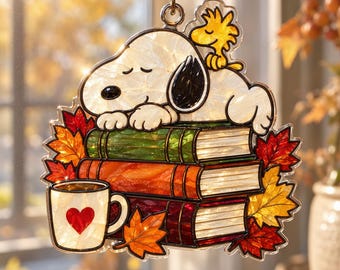 May include: A stained glass suncatcher featuring Snoopy sleeping on a stack of three books with Woodstock perched on top. The design includes a white mug with a red heart and colorful autumn maple leaves in shades of orange, red, and yellow.