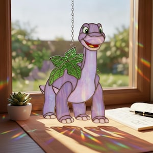 Könnte beinhalten: Ein Buntglas-Suncatcher in Form eines hellvioletten Dinosauriers mit einem grünen Blatt auf dem Rücken. Der Dinosaurier hat einen langen Hals, ein lächelndes Gesicht mit großen grünen Augen und hängt an einer silbernen Metallkette vor einem Fenster.
