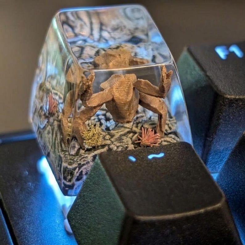May include: A clear resin mechanical keyboard keycap featuring an encapsulated brown crab figure resting on a textured seabed with small red and yellow coral-like accents. The keycap is positioned on a black keyboard next to a key with a tilde symbol.