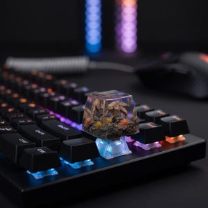 May include: A clear resin mechanical keyboard keycap featuring a miniature brown creature sculpture, textured rock base, and small orange and yellow tufts of grass, installed on a black keyboard with illuminated blue and orange light switches.