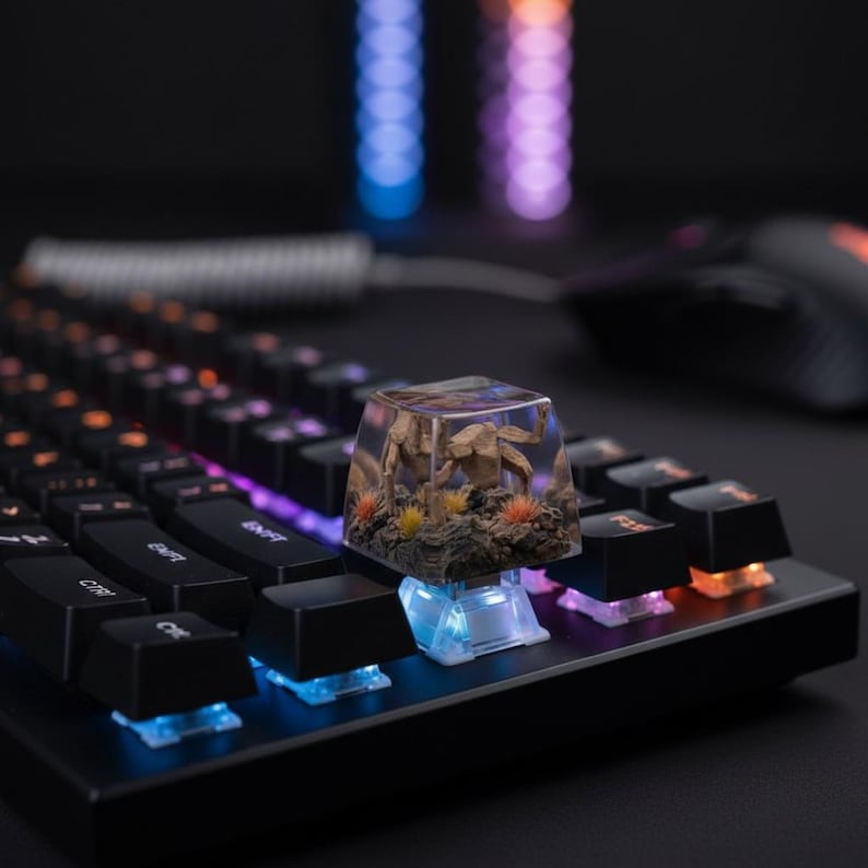 May include: A clear resin mechanical keyboard keycap featuring a miniature brown creature sculpture, textured rock base, and small orange and yellow tufts of grass, installed on a black keyboard with illuminated blue and orange light switches.