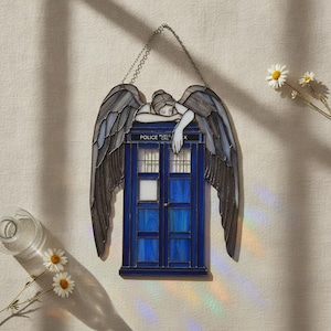 May include: A stained glass suncatcher featuring a blue police box with the text POLICE CALL BOX, topped by a figure with large, dark, textured wings resting their head on the roof. The piece is suspended by a metal chain for display.