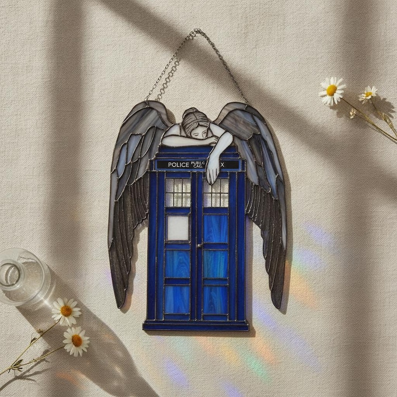 May include: A stained glass suncatcher featuring a blue police box with the text POLICE CALL BOX, topped by a figure with large, dark, textured wings resting their head on the roof. The piece is suspended by a metal chain for display.