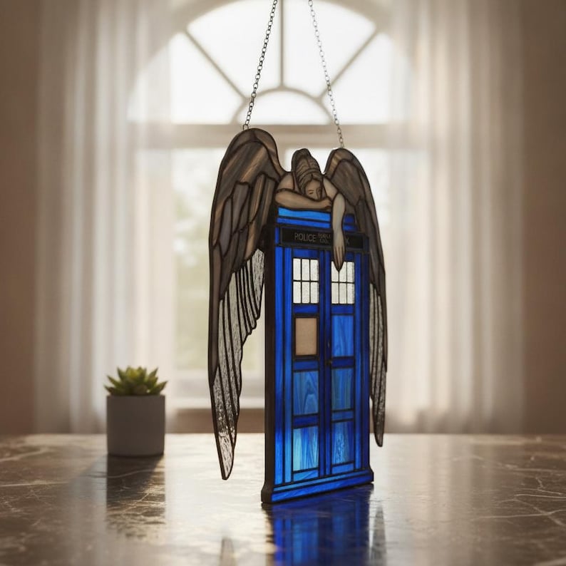 May include: A stained glass hanging ornament featuring a blue police box with the text POLICE BOX visible above the door. A grey winged figure with detailed feathers rests its arms on top of the box. The piece is suspended by a metal chain.