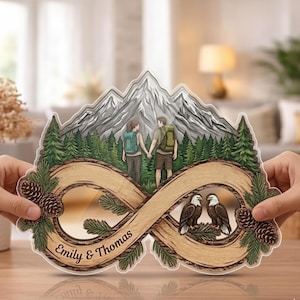 May include: A wooden-style infinity sign with a mountain and forest scene, featuring a couple hiking and two eagles. The sign includes the names "Emily & Thomas" and is decorated with pinecones and evergreen branches.