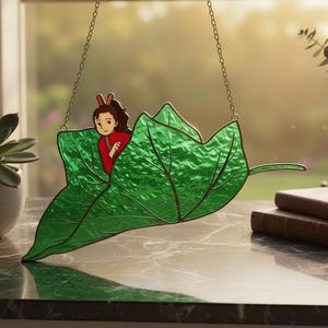 Op de afbeelding: Een glas-in-lood suncatcher met een groot, getextureerd groen blad met opvallende nerven. Een kleine, geïllustreerde figuur met donker haar en rode kleding is achter het blad geplaatst. Het object hangt aan een dunne metalen ketting.