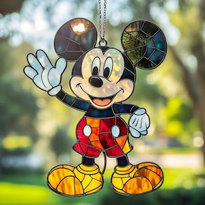 Op de afbeelding: Een glas-in-lood suncatcher in de vorm van Mickey Mouse, met zwarte oren, een wit gezicht, een rode broek met gele knopen en gele schoenen. Het object is gemaakt met donkere loden lijnen en hangt aan een dunne, zilverkleurige metalen ketting.