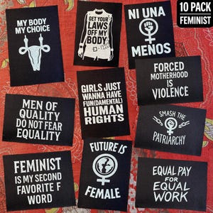 Peut inclure: Dix écussons noirs avec des inscriptions féministes blanches. On peut lire des phrases comme « Mon corps, mon choix », « Briser le patriarcat » et « À travail égal, salaire égal ». Les écussons sont disposés sur un fond rouge à motifs.