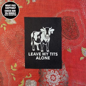 Könnte beinhalten: Ein schwarzer Aufnäher mit einer weißen Kuh-Illustration und dem Text "LEAVE MY TITS ALONE". Der Aufnäher hat auch eine runde Grafik mit dem Text "WANT FREE SHIPPING? CHECK OUR 10x BUNDLE LINK IN DESCRIPTION".