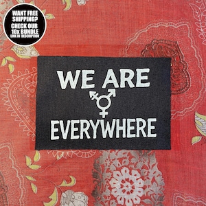 Op de afbeelding: Een zwarte rechthoekige patch met de woorden "WE ARE EVERYWHERE" in witte blokletters. Een symbool dat de transgender identiteit vertegenwoordigt staat in het midden. De patch ligt op een rode stof met een patroon.