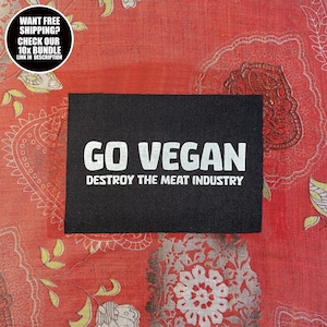 Może przedstawiać: Czarna prostokątna naszywka z napisem "GO VEGAN" dużymi białymi literami nad "DESTROY THE MEAT INDUSTRY". Naszywka jest umieszczona na czerwonej tkaninie we wzory. Okrągła grafika w lewym górnym rogu głosi "WANT FREE SHIPPING? CHECK OUR 10X BUNDLE LINK IN DESCRIPTION."
